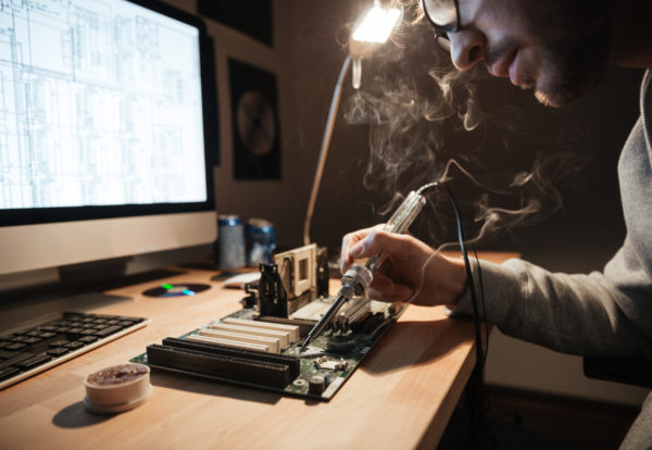 Closeup of serious young man using scheme on monitor and soldering iron for repairing motheboard